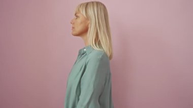 Orta yaşlı bir kadın, kendine güveni tam bir sarışın gömlek giyiyor ve pembe bir arka planda doğal bir şekilde gülümsüyor.