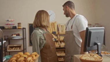 Man and woman bakers in a shop talking with shelves of bread and pastries around them in an indoor bakery setting