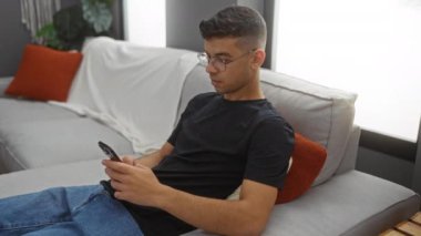 Genç adam modern dairede koltukta dinleniyor oturma odasındaki telefona bakıyor rahat ve rahat bir ev ortamı öneriyor.