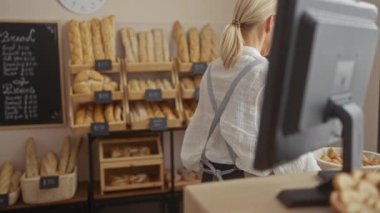 Fırında çalışan sarışın kadın müşterilere servis yapıyor ve ev içi raflarda ekmek düzenliyor.