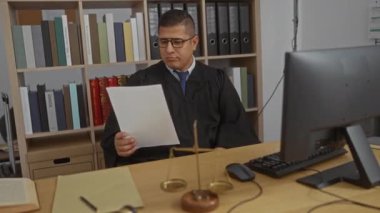 Gözlüklü ve yargı cüppeli genç adam, arka planda bilgisayar, adalet terazisi ve kitaplık olan bir ofiste belge okuyor.