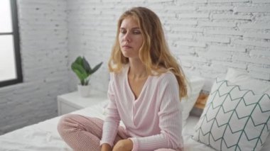 Genç bir kadın beyaz tuğla duvarlı, parlak bir yatak odasında oturuyor. Rahat pembe pijamalarını giyerken dalgın ya da endişeli görünüyor.