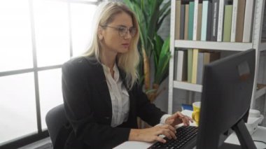 Ofiste bilgisayar başında çalışan, gözlük takan, klavyeye yazı yazan, kitaplarla çevrili, sarı bardak tutan, sarı saçlı, profesyonel kıyafetli bir kadın..