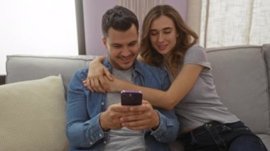 Oturma odalarında birlikte gülümseyen ve telefona bakan çift kapalı mekanda mutlu bir ilişki sergiliyor..