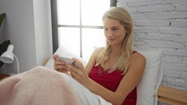 Yatakta akıllı telefon kullanan kadın gülümsüyor. Rahat bir şekilde sabah güneşinin tadını çıkarıyor.