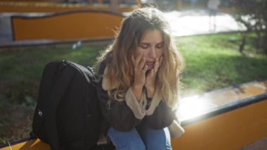 Bir bankta oturan kadın, elleri güneş parkında endişeli bir şekilde oturuyor. Sırtına siyah bir sırt çantası takıyor..