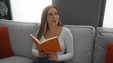 Modern bir oturma odasında rahat bir koltukta kitap okuyan bir kadın, bir rahatlama anı yakalıyor ve iç mekan gözlemi yapıyor..