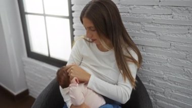 Anne, bebeğini rahat oturma odasında emziriyor. Yumuşak doğal ışıklandırmayla, modern ev dekorlarıyla çevrili, kadın ve kız arasında sıcaklık ve bakım taşıyor..