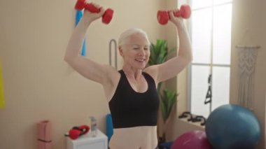 Yaşlı kadın spor salonunda ağırlıklarla egzersiz yapıyor, kapalı bir spor merkezinde dayanıklılık ve zindelik gösteriyor..