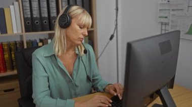 Ofiste çalışan, kulaklık takan ve bilgisayar kullanan olgun bir kadın arka planda kitap ve klasörlerle dolu bir masada oturuyor.