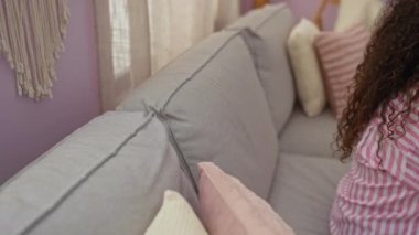 Kanepede uzanmış kıvırcık saçlı, dekoratif yastıklı ve makrome duvar resimli genç bir kadın.