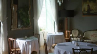 Yuvarlak masaları, beyaz masa örtüleri, sandalyeleri ve perdeleri olan büyük pencerelerden süzülen güneş ışığı olan şık bir kapalı restoran manzarası.