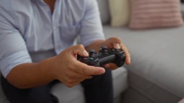 Evde video oyunu oynayan bir adam oturma odasında gri bir kanepede otururken mavi gömlek giyen bir oyun kumandası kullanıyor.