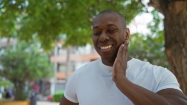 Beyaz gömlekli genç adam, yeşil ve bulanık şehir arka planıyla çevrili güneşli bir şehir parkında çenesini tutarken gülümsüyor..