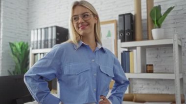 Modern ofis ortamında güvenle gülümseyen sarı saçlı genç bir kadın. Gözlük takıyor ve mavi gömlekli. Etrafı kitaplık ve yeşil bitkilerle çevrili..