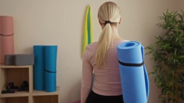 Spor salonunda gülümseyen bir kadın, yoga minderi, sarı saç, pembe gömlek, arka planda bitkiler, fitness ekipmanları, kapalı ortamlar, hoş geldin jestleri, spor merkezi atmosferi..