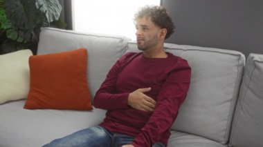 Evdeki kanepede oturan genç adam, sıcak bir oturma odasında kederli bir ifadeyle karnını tutuyor..