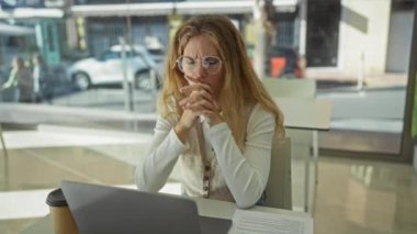 Ofiste dizüstü bilgisayarlı, kahve bardaklı, günlük kıyafetler giyen, kapalı mekanlı, düşünceli, düşünceli, profesyonel bir çevrede çalışan bir kadın..