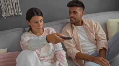 Çift evde bir kanepede oturup, tarafsız iç dekorasyonlu bir oturma odasında televizyon izliyor.