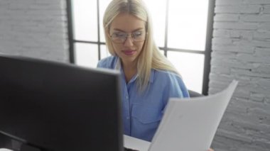 Modern ofiste bilgisayar başında çalışan bir kadın, geniş pencerelerin ışığı altında mavi gömlek ve gözlük takan, odaklanmış ifadeli belgeleri inceliyor..