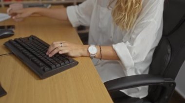 Masada çalışan, klavyede yazan, ofis odasında telefonla konuşan, etrafı kitap ve dosyalarla çevrili, beyaz gömlek ve gözlüklü, ellerine odaklanmış, sarı saçlı bir kadın.