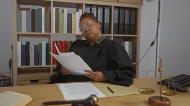 Siyah cüppeli kadın yargıç büro masasında tokmak pulları ve arkaplandaki dosyaları okuyor..