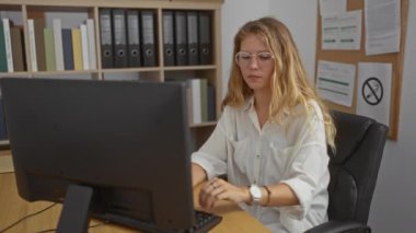 Ofisteki bir masada çalışan bir kadın, elinde bir tablet, elinde klasörler ve kitaplarla bilgisayar kullanıyor.