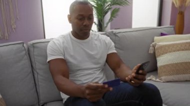 Kapalı bir yerde akıllı telefon ve kredi kartı tutan bir adam, rahat oturma odasında, tarafsız bir dekorla, düşünceli bir ifade sergiliyor, modern apartmandaki gri koltukta rahat bir şekilde oturuyor..