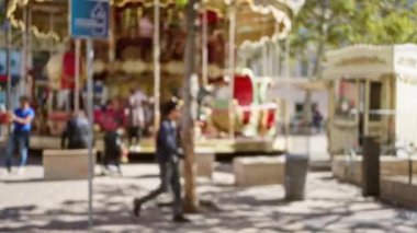 Marsilya 'daki bulanık ağaçlar ve oyun alanının arka planında güneşli bir caddede atlıkarıncanın yanında yürüyen insanları gösteren odaksız bir sahne..