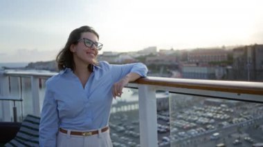 Gezgin gemi rıhtımında gülümseyen bir kadın, şehir siluetine dayanan gözlükleriyle, günlük mavi gömlek giymiş, günbatımının tadını çıkaran, genç zarafeti ve açık havada huzurlu mutluluğu somutlaştıran bir kadın..