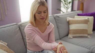 Oturma odasındaki kanepede oturan pembe gömlekli bir kadın endişeli görünüyor, bileğini tutuyor, rahat bir evde bilek ağrısı ya da rahatsızlık hissi veriyor..