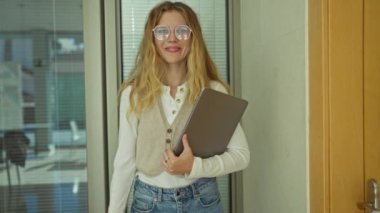 Ofiste el kol hareketi yapan, gülümseyen, dizüstü bilgisayarı tutan, kameraya bakan, yelek ve gözlük takan, sarı saçlı, kapının yanında duran, kapalı alan ayarlı, iş atmosferi olan bir kadın..