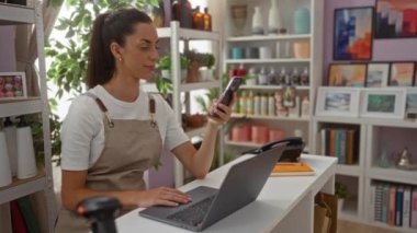 Ev dekorasyonu mağazasında çalışan genç bir kadın, elinde telefon ve dizüstü bilgisayarla etrafı düzgün düzenlenmiş ürünler ve dekorasyonlarla çevrili bir tezgahta çalışıyor.