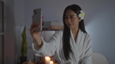 Spa 'da akıllı telefonuyla selfie çeken bir kadın. Saçında çiçekli beyaz bir bornoz ve gevşetici bir güzellik merkezinde mumlarla çevrili bir kadın.