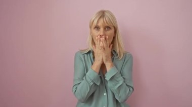 Orta yaşlı bir kadın, sarışın, tişört giymiş, şokta, elleri ile ağzını kapatıyor, pembe arka planda bir hata yaptıktan sonra, bir sır saklıyor, ifadesinden utanıyor.