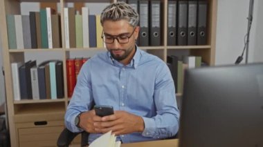 Mavi gömlekli ve gözlüklü genç adam modern ofis odasında kitaplık ve klasörlü akıllı telefon kullanıyor.