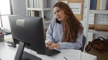 Ofiste çalışan bir kadın telefonla konuşuyor ve masasında bilgisayar ve belgelerle kitap rafları ve iş aksesuarlarıyla çevrili profesyonel bir iş yerinde notlar yazıyor..