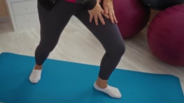 Spor salonunda yoga minderinin üzerinde spor kıyafetleri giymiş spor topları ve spor aletleri ile çevrili bir kadın içeride sağlık ve sağlığa odaklandığını gösteriyor..