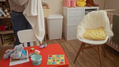 Bebek odasını düzenleyen kadın oyuncak ve battaniyelerle dolu çamaşır sepetini tutuyor. Doldurulmuş hayvanlar ve renkli dekorlarla çevrili. Sıcak ve neşeli bir ortam yaratıyor..