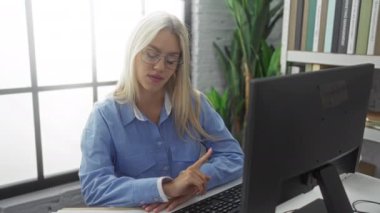 Modern ofiste bilgisayar başında konuşan bir kadın. Etrafı kitaplarla çevrili. Mavi tişört, sarı saç, gözlük, camdan gelen doğal ışık..