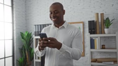 Ofiste gülümseyen genç adam pencerenin yanındaki akıllı telefonu kullanıyor. Arka planda bitkiler var. Parlak, modern çalışma alanı ayarlarını gösteriyor..