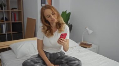 Yatak odasında, rahat bir odada, akıllı telefonlu, gülümseyen esmer bir kadınla selfie çeken bir kadın. Evde günlük kıyafetlerle oturuyor. Keyifli bir yüz ifadesi sergiliyor.