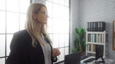 Kadın, modern ofiste güven içinde poz veriyor, gözlük takıyor ve iş kıyafetleri giyiyor, parlak pencereleri ve bitkileri olan, profesyonel, misafirperver bir ortam yaratıyor..