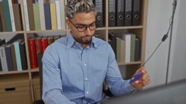 Ofiste oturmuş, bilgisayarında çalışırken ve kitaplarla ve dosyalarla çevrili gözlüklü genç bir adam.