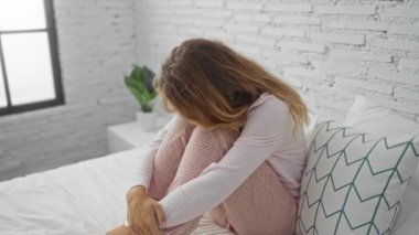 Beyaz tuğladan bir yatak odasında oturan, üzgün ve düşünceli, rahat pijama giysileri giyen, sarı saçlı, kısmen iç mekanda, ev ortamında oturan bir kadın.