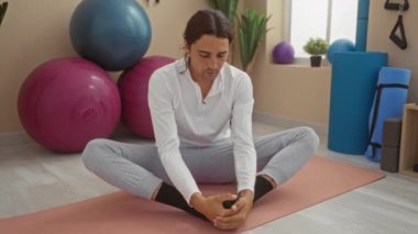 Uzun saçlı, beyaz tişörtlü ve gri pantolonlu bir adam spor salonunda yoga minderine uzanmış spor topları ve spor aletleriyle çevrili.