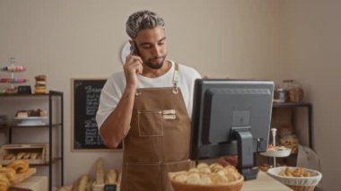 Pastanede çalışan sakallı genç bir adam. Kapalı mekanda telefonla konuşurken bilgisayar kullanıyor.