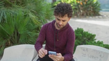 Parkta oturan genç bir adam, eli yüzü üzerinde, yeşillikle çevrili, bordo kapüşonlu kapüşonlu, şehir dışında bir bankta açık hava sahnesi giymiş, stresli görünüyor..