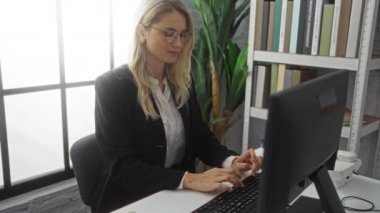 Bilgisayarda çalışan bir kadın, sarı saç, gözlük, arka planda kitaplık ve bitki bulunan belgelere odaklanmış, profesyonel bir iş ortamı yaratıyor..