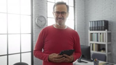 Elinde tablet olan kırmızı kazaklı olgun bir adam modern ofiste el sıkışmak için uzanır. Kitaplıklar ve büyük pencereler ile..